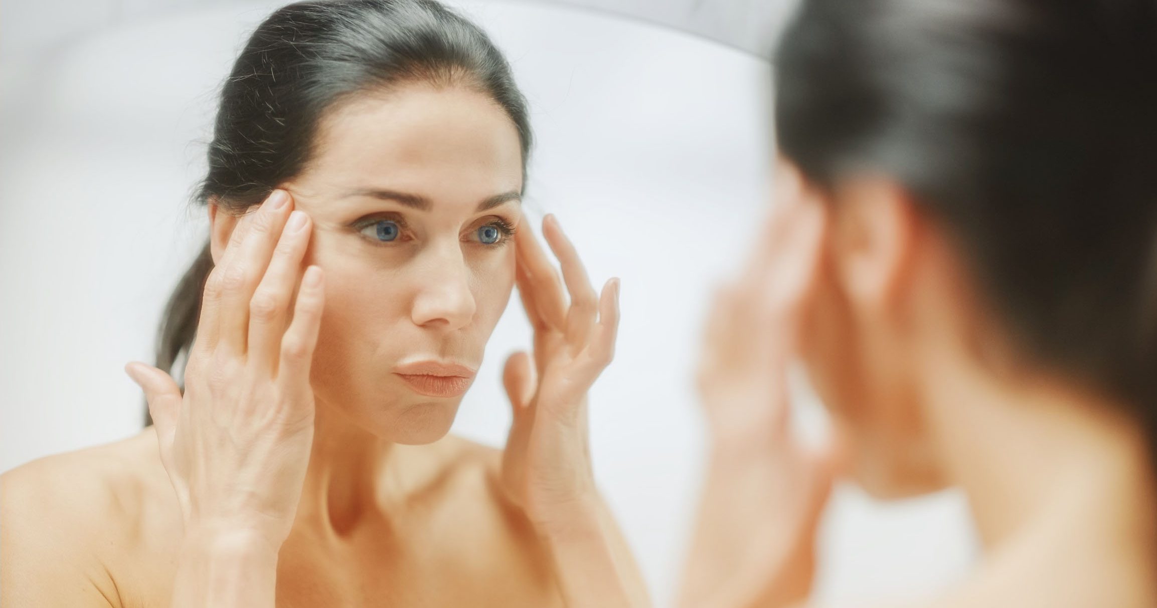 A woman in her 40s looking at herself in a mirror and thinking about facelift surgery.