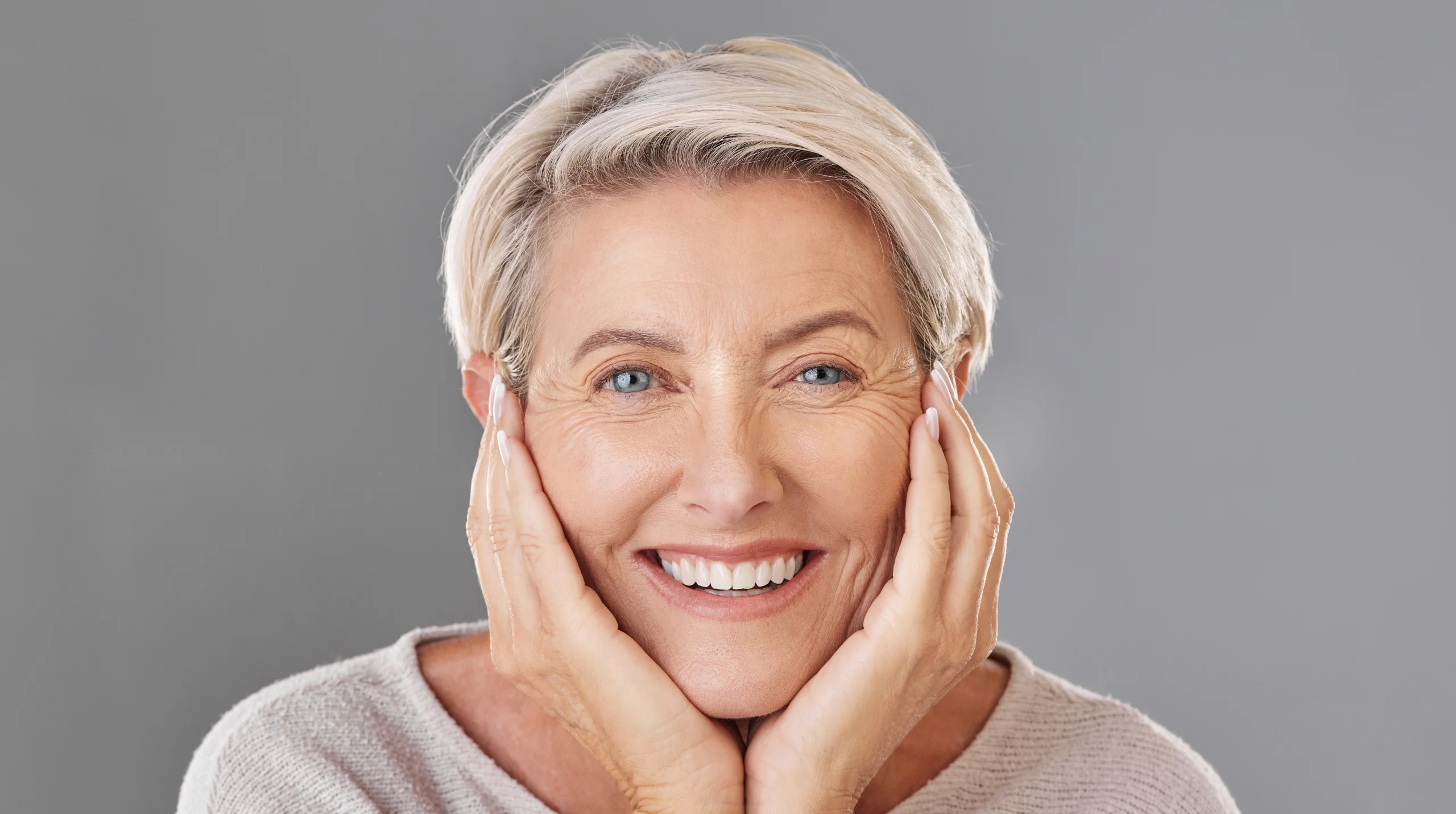 front facial shot of beautiful mature woman. Her hands are cupping under her chin, and she is smiling.